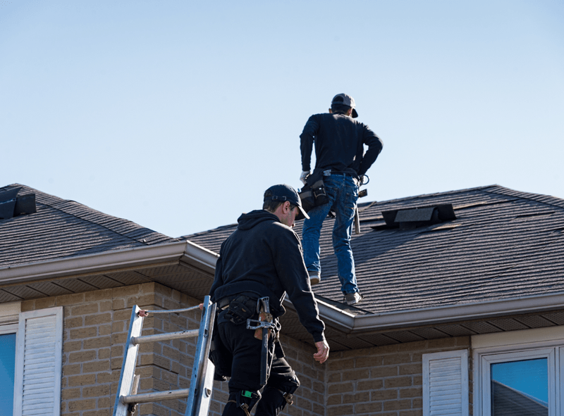 two roofing contractors wearing black standing on roof with tool belts next to ladder