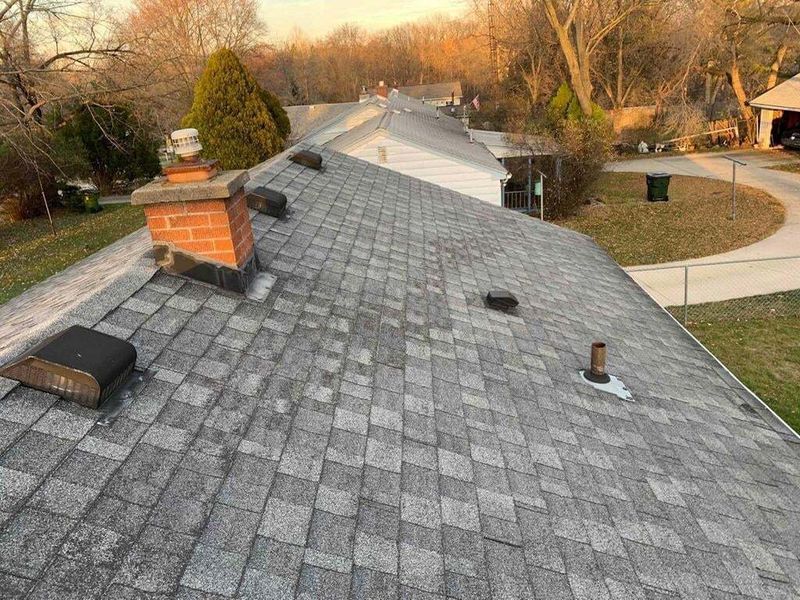old roof with black and gray shingles red brick chimney and roof vent trees in background